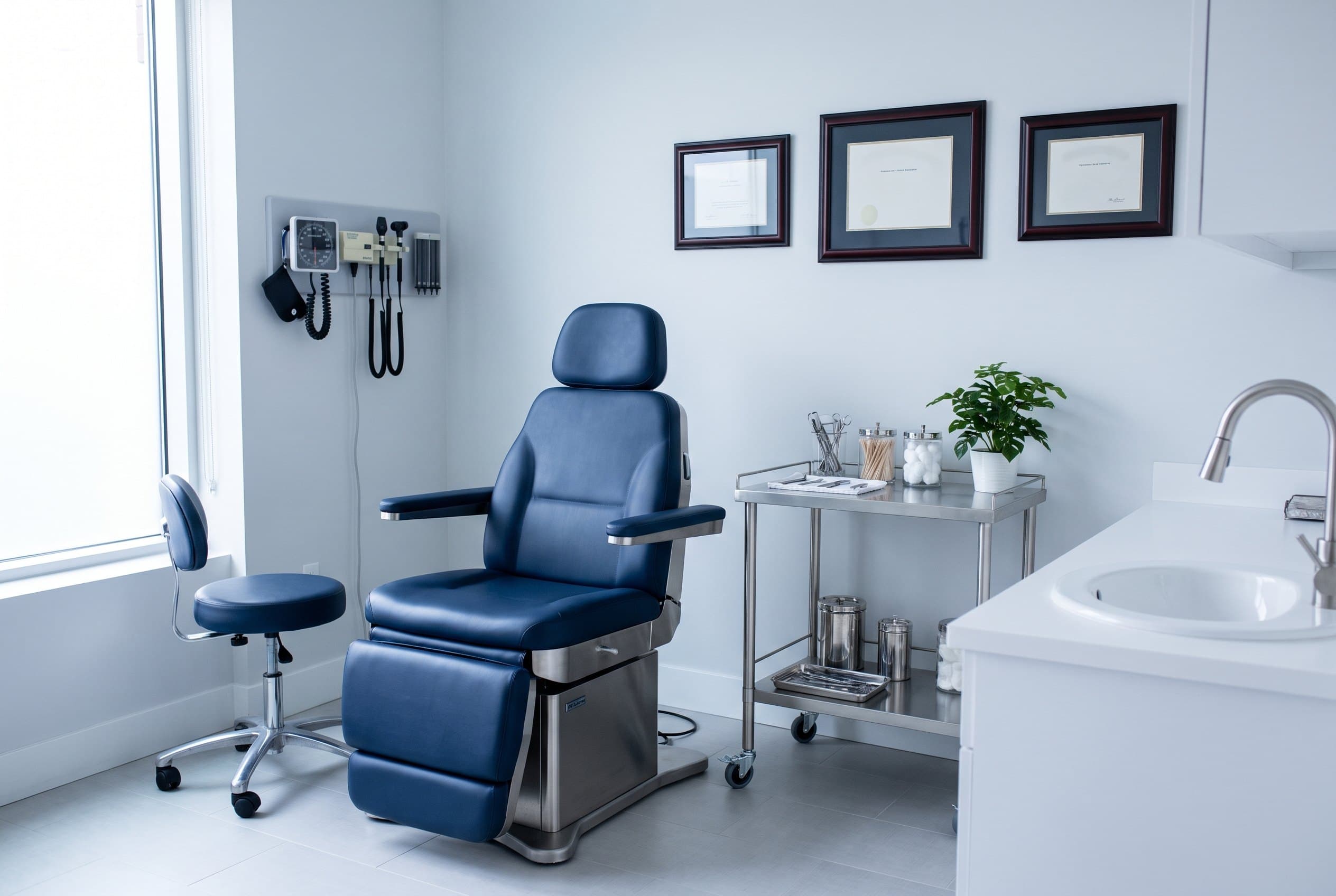 A treatment room with a navy medical chair, wall-mounted instruments, a steel trolley and framed diplomas